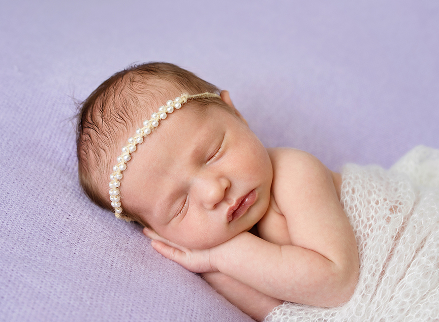 Newborn baby girl with pearl headband on purple wrapped in cream in a studio portrait